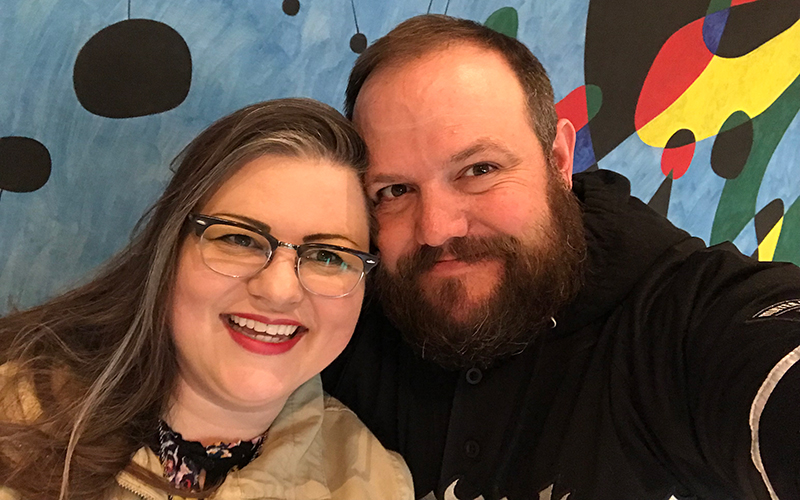 Two white people smile in front of a mural