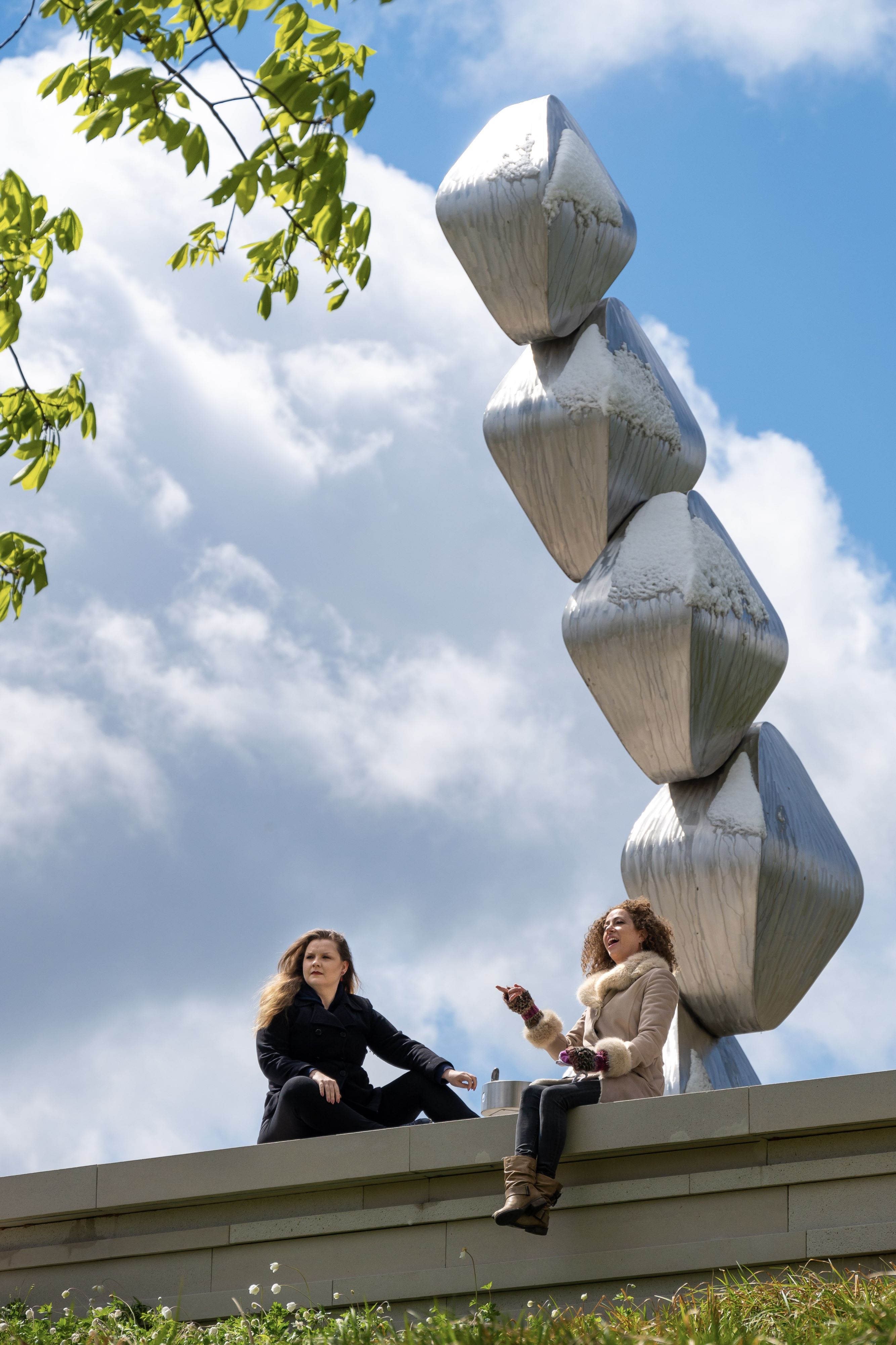 Two women sit on a stone wall with a metallic sculpture in the background.