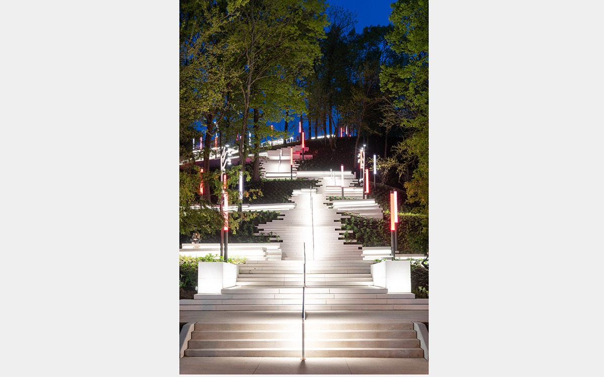 An outdoor staircase, lit up at night, climbs up a tree covered hill.