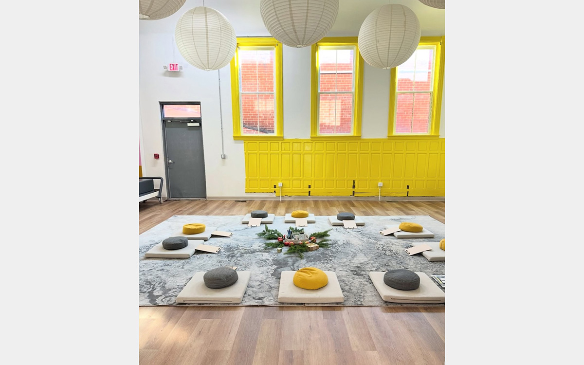 10 meditation cushions on a gray carpet surround a flower arrangement; behind is a yellow wall with yellow-framed windows; above are round paper lanterns