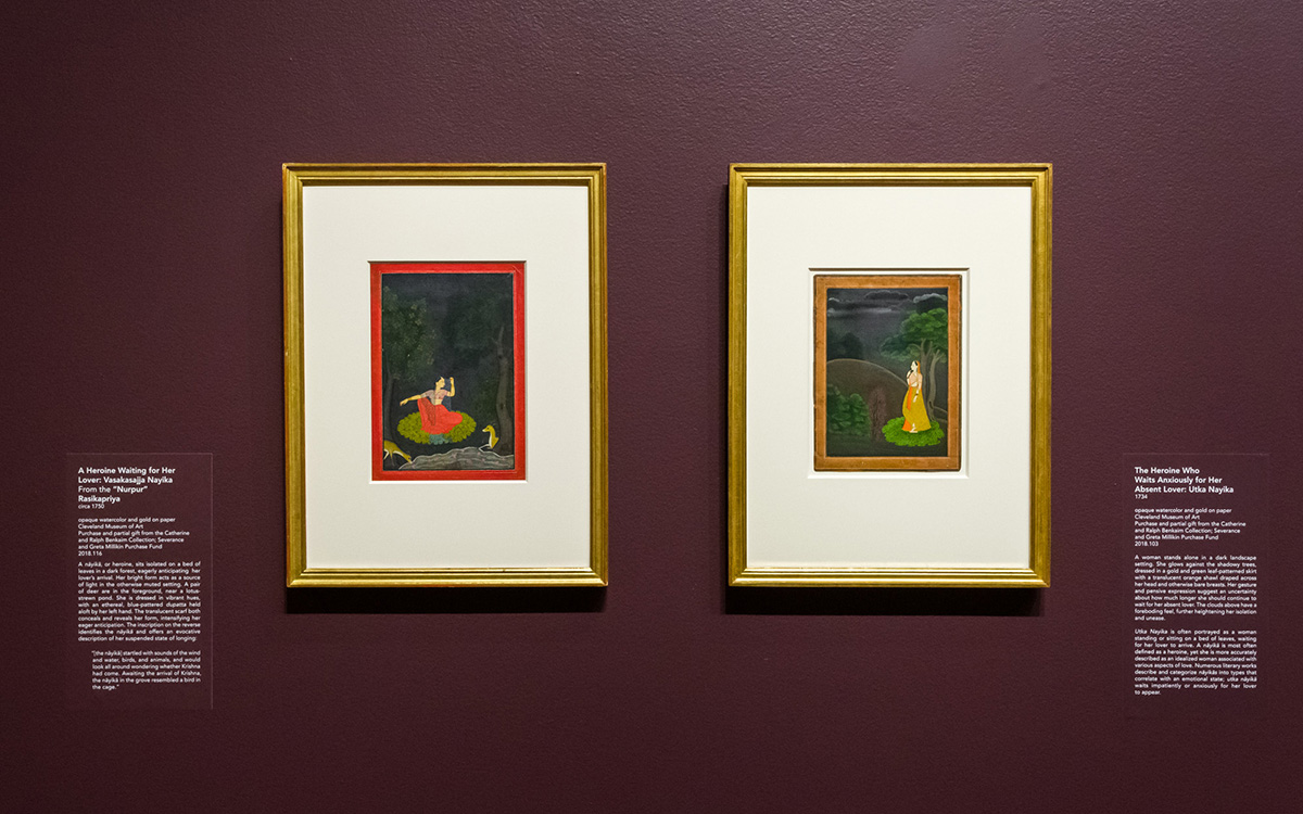 Two colorful framed paintings hang on a burgundy-colored wall with interpretive labels in white lettering on either side of the photo..