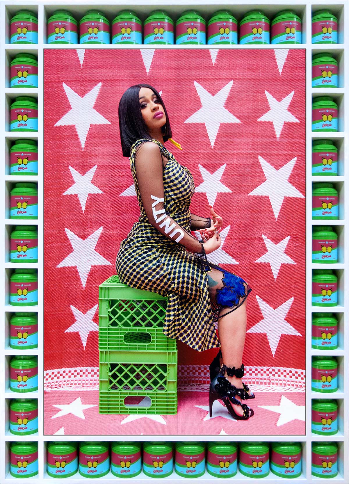 Cardi B sits on two green milk crates and looks at the camera. The word "unity" is spelled out on her arm. The image is surrounded by a border made of bright green, blue, and pink tea boxes.