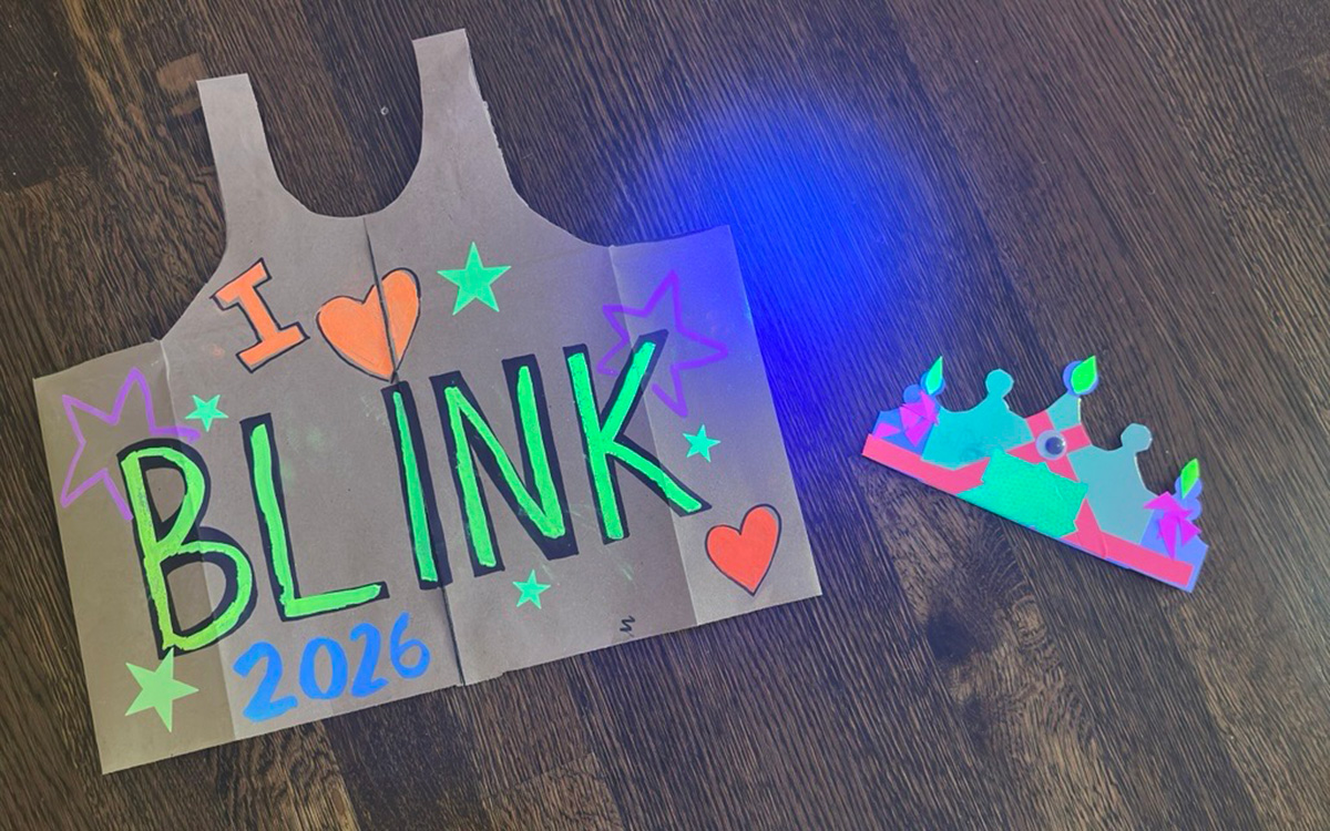 Paper vest and crown decorated with fluorescent paints against a wood flooring background. "I heart BLINK 2026" is written on the vest.