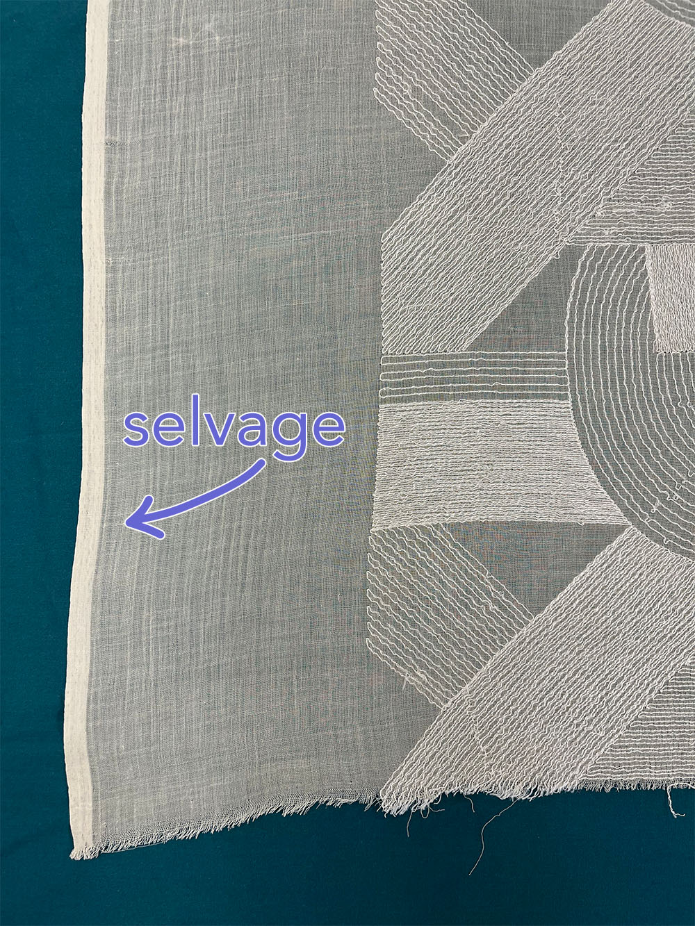 Close-up of a light-colored fabric with a geometric pattern including rectangles & semicircles. The thicker selvage and the ragged, raw edge are both visible.