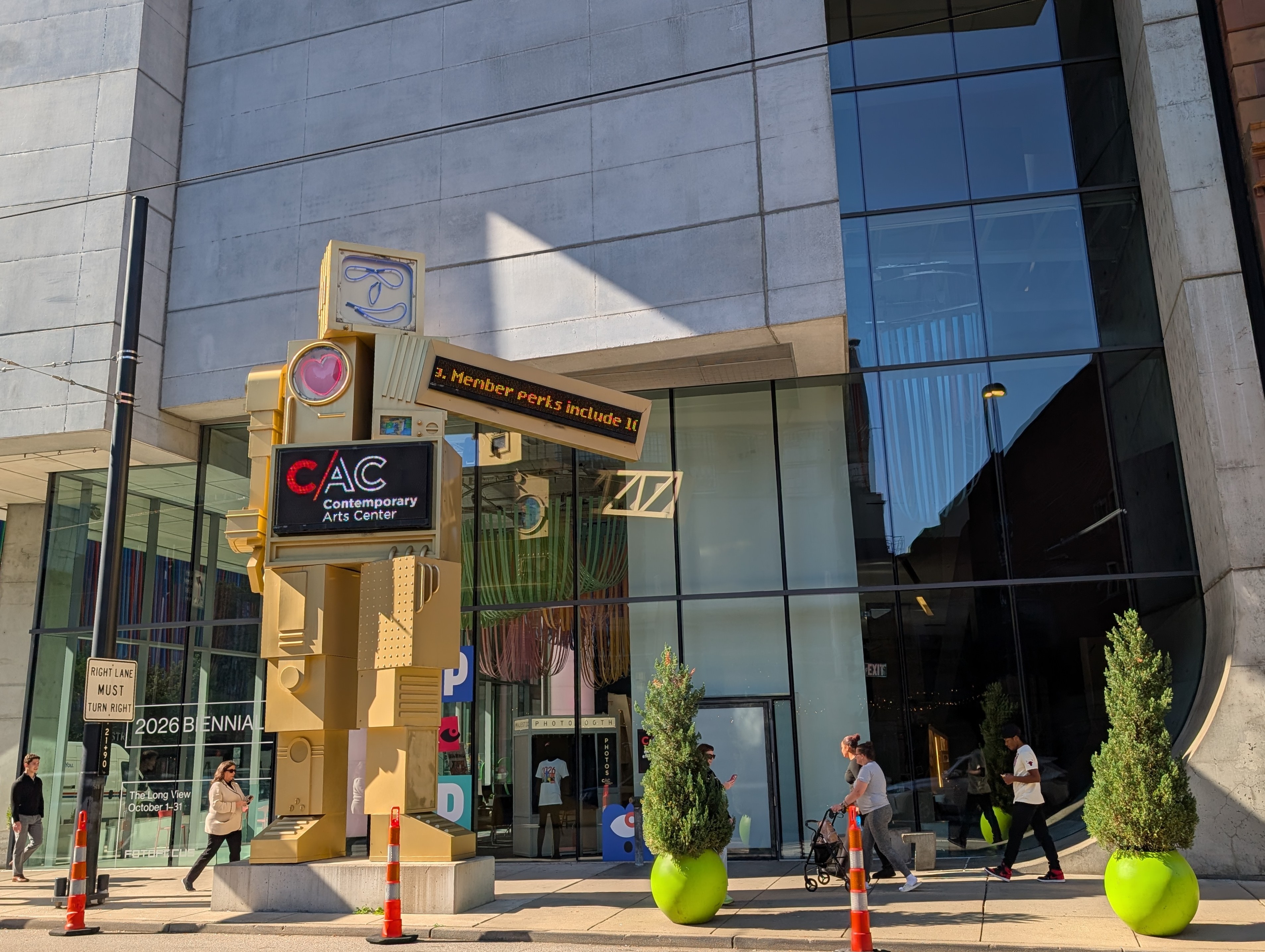 Large robot-like outdoor sculpture on a sidewalk in front of a concrete and glass building with people walking past.