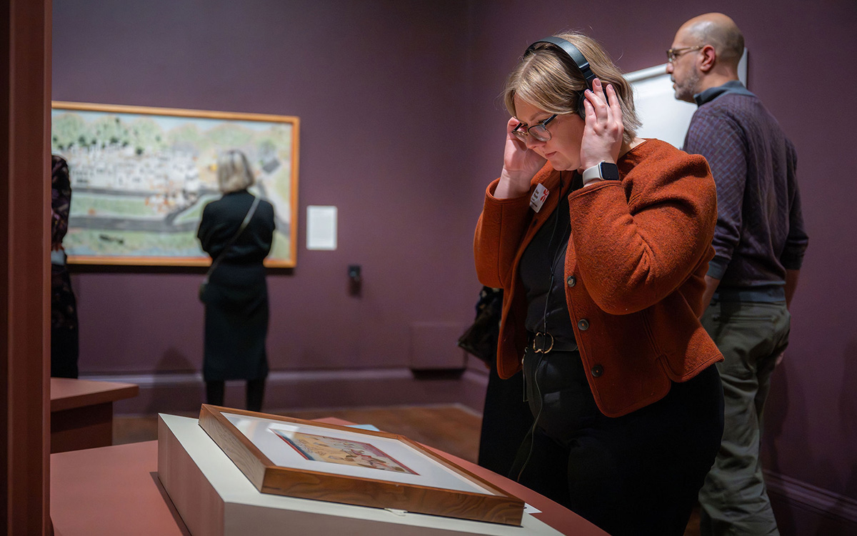 A woman wears headphones while looking at a framed artwork sitting on a display table while people in the background look at framed artwork hanging on the walls.