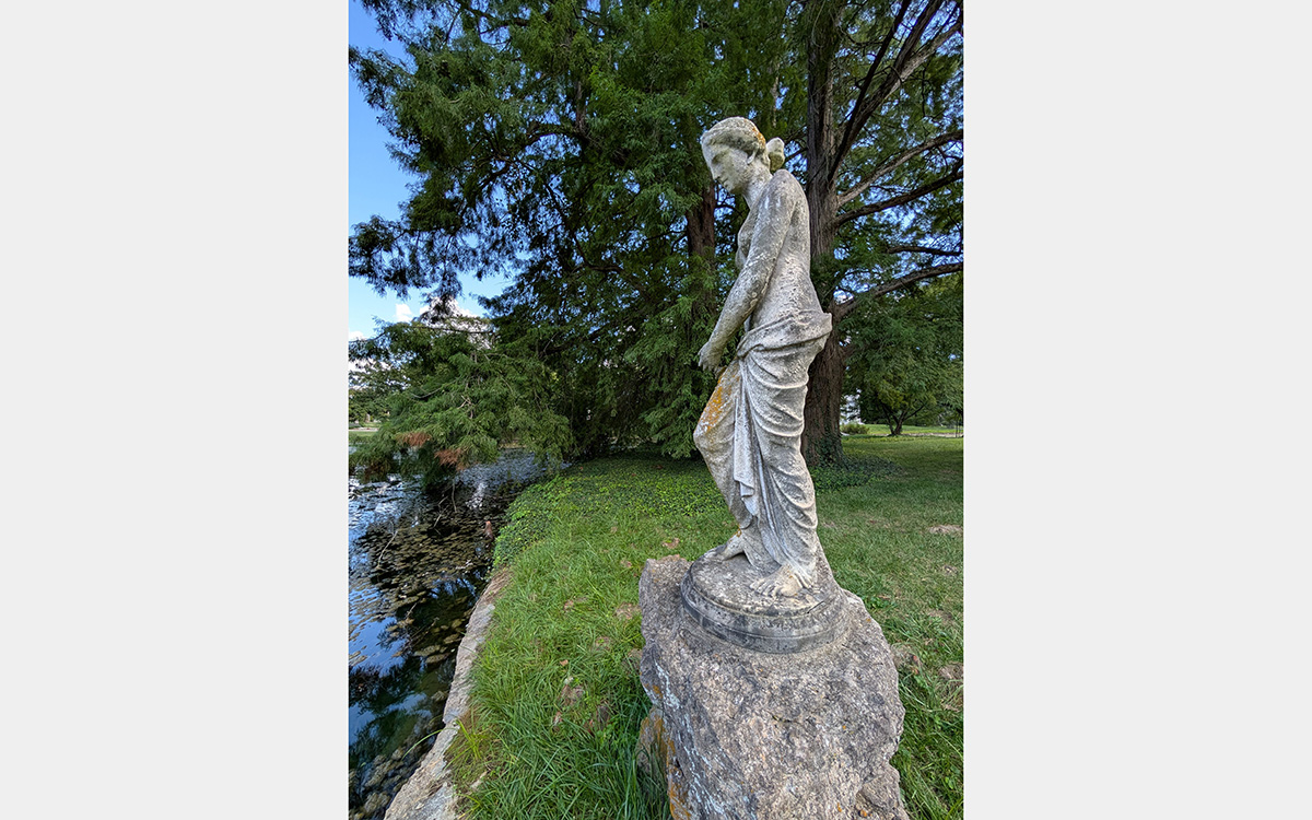 A statue of a woman stands on a base of rough-hewn rocks next a pond in a green park-like setting.