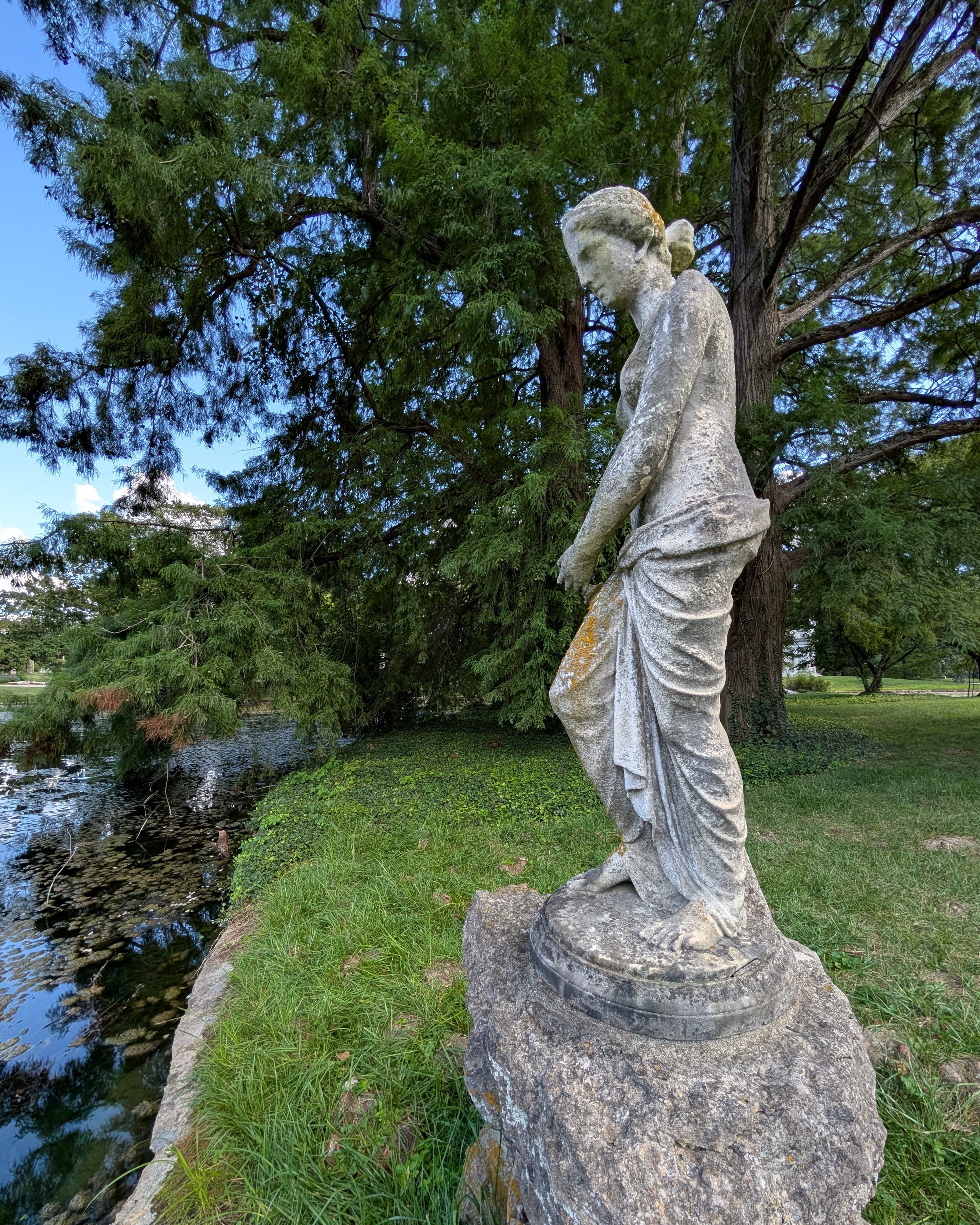 A statue of a woman stands on a base of rough-hewn rocks next a pond in a green park-like setting.