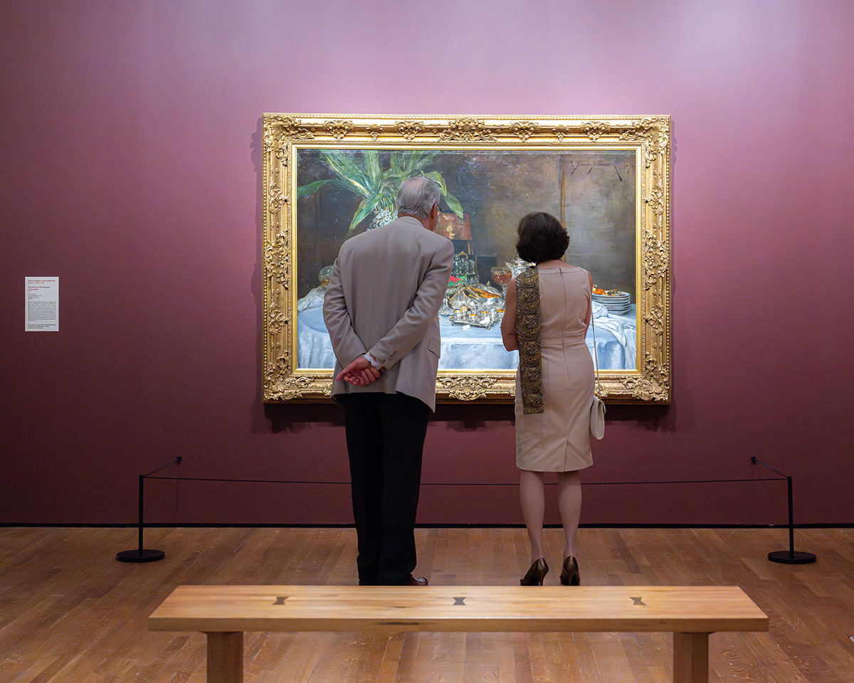Two people with their backs to the camera view a large painting in a gold frame against a purple wall. A wood bench stands in the foreground.