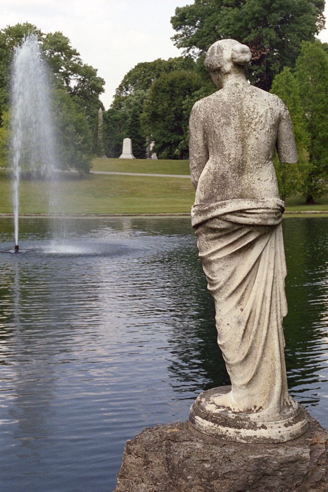 Back of a marble statue of a woman overlooking a lake with a fountain and green landscape in the background.