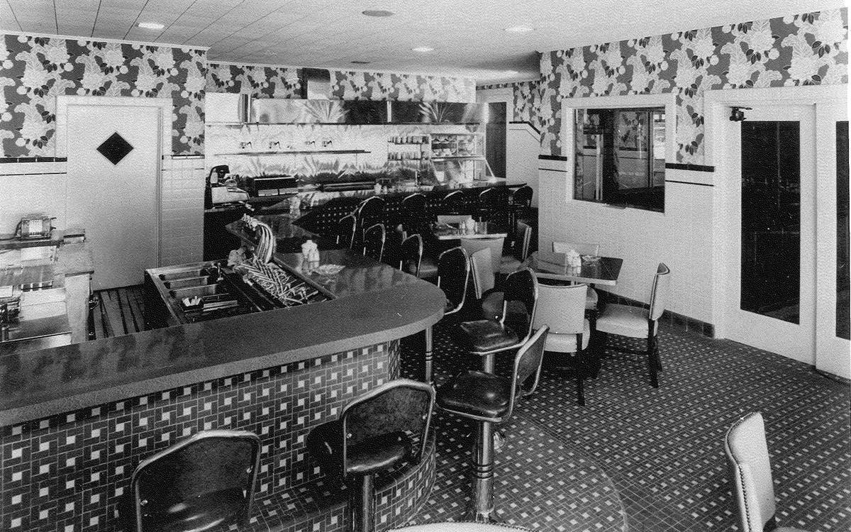 Black and white photo of a dining counter inside a restaurant with patterned carpets, tiles, and wallpaper