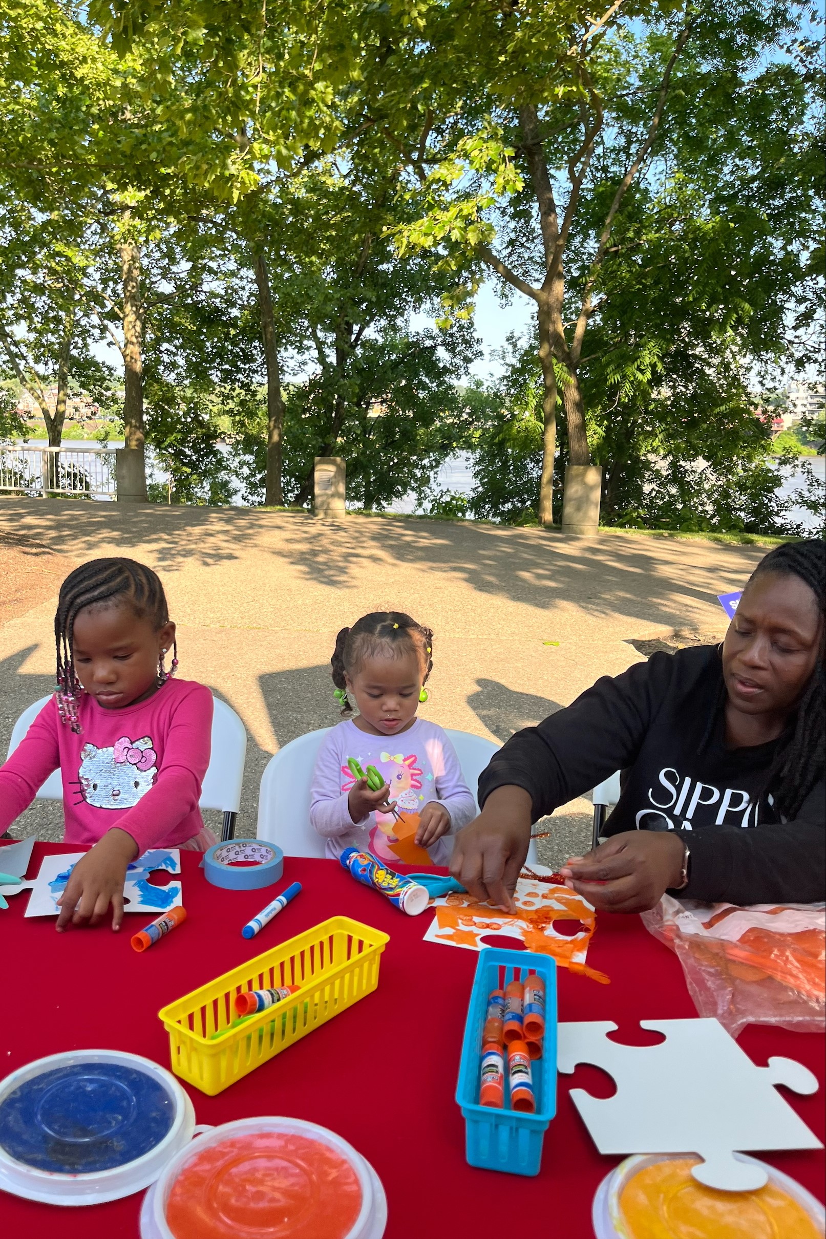 Two girls and woman make art on a table with trees and a body of water in the background.