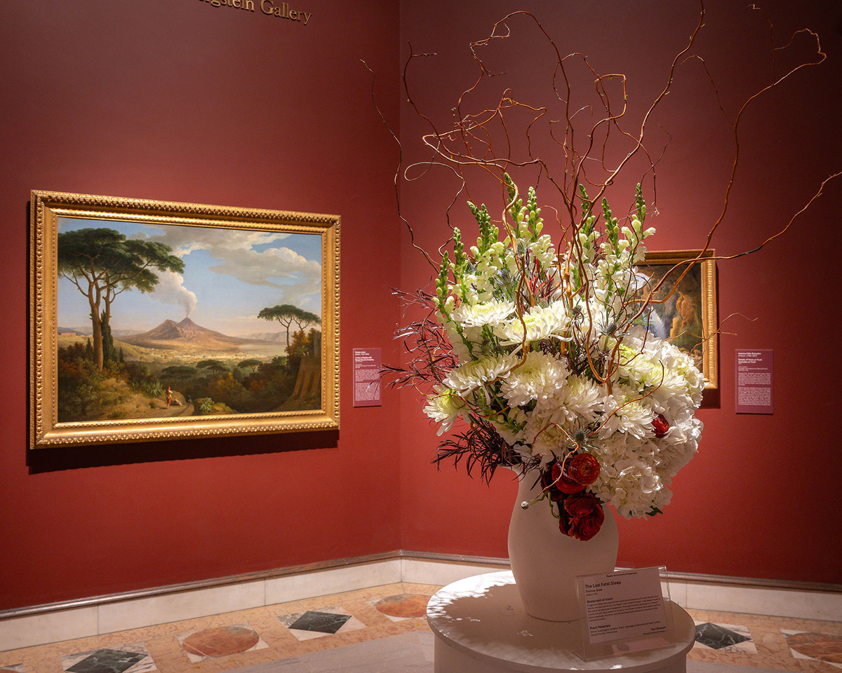 A large white vase contains a flower arrangement with large white flowers and few red roses inside a room painted a bright orange-red. A painting of a landscape with a volcano inside a gold frame hangs in the background. 