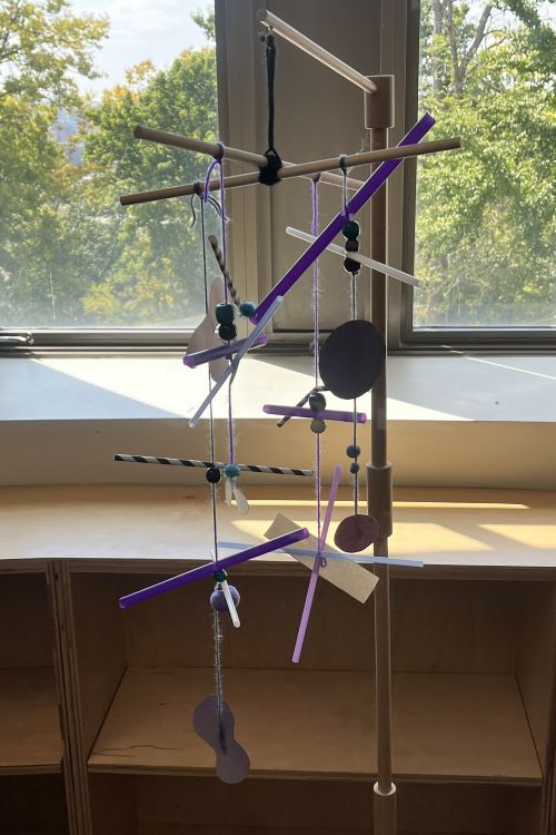 Mobile made of colorful straws and cut out shape against a background of wooden shelves and windows looking out on green trees.