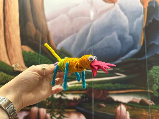 A human hand holds an animal-like figure made of yard against the backdrop of a landscape painting.