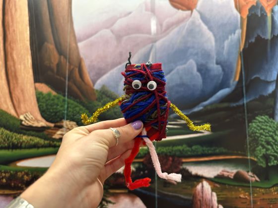 A human hand holds an animal-like figure made of yard against the backdrop of a landscape painting.