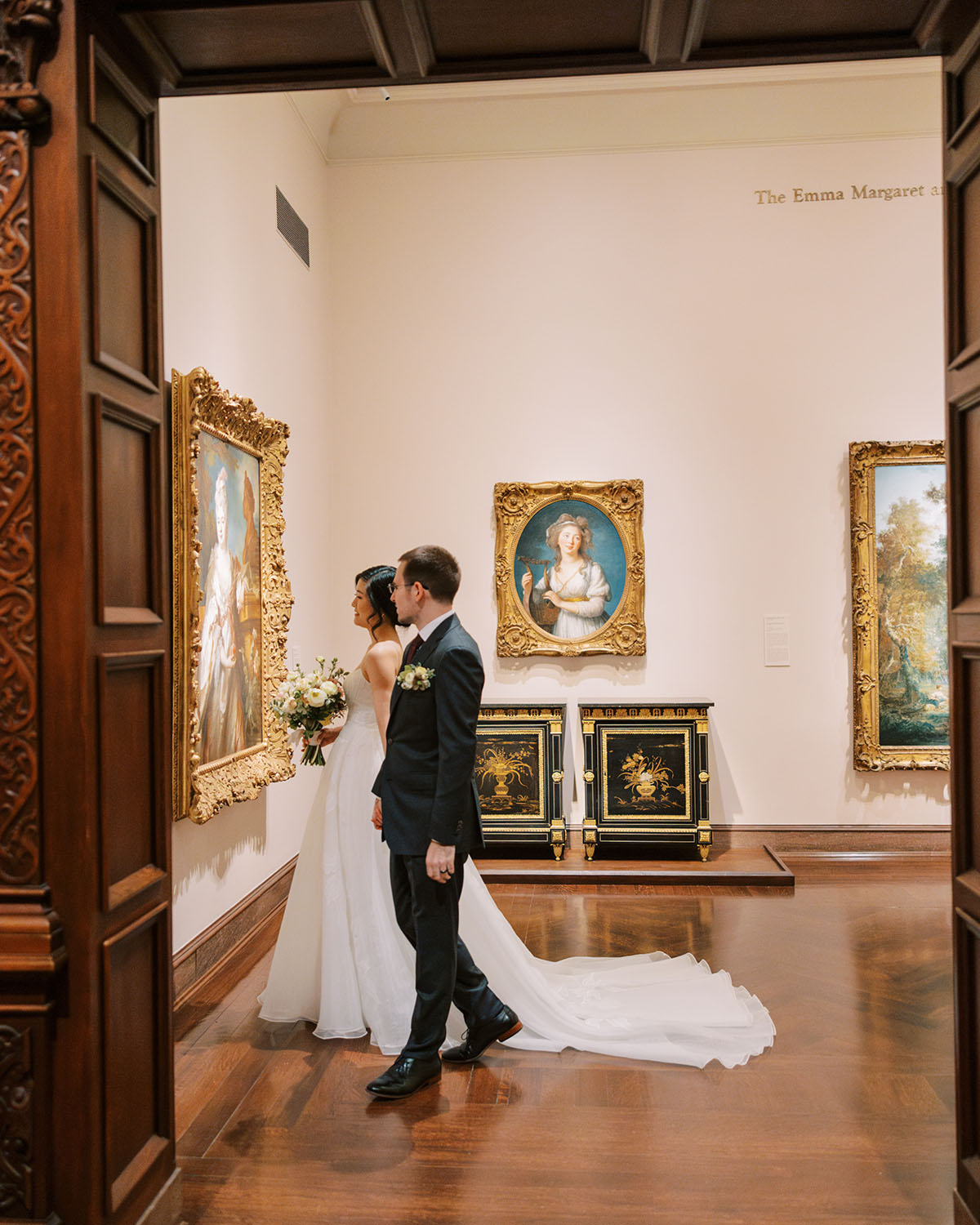 The couple look at a painting in an ornate, golden frame