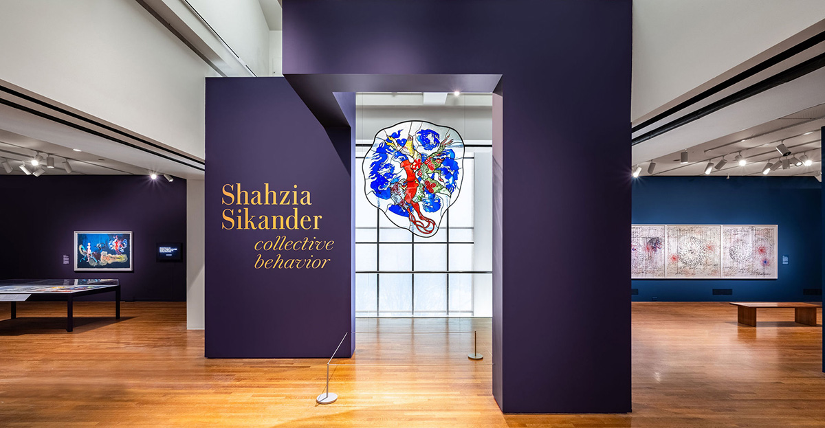 A large red and blue glass piece hangs from a purple wall that says "Shahzia Sikander: Collective Behavior"