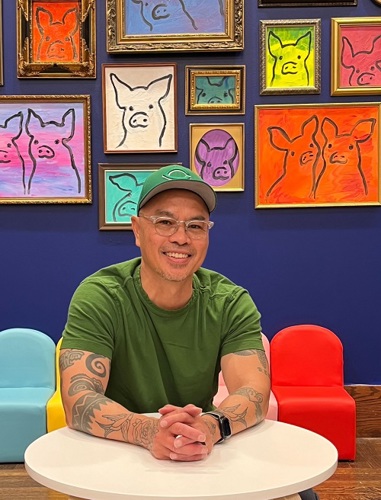 Artist John Lanzador wearing a green hat and green shirt in front of a vibrant display of framed art featuring pigs.