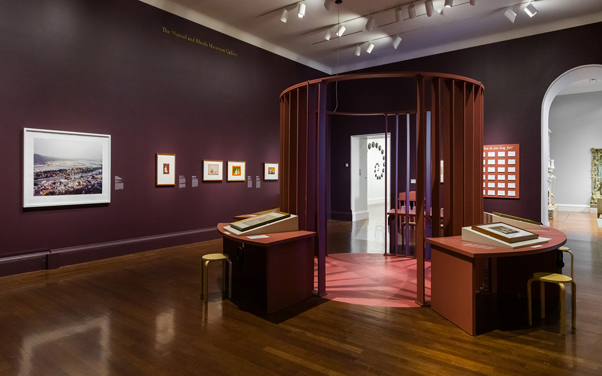 Circular structure made of vertical slats sits in the middle of an exhibition space surrounded by tabletop displays and framed artwork hung on walls.