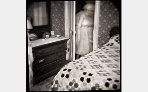 Nancy Rexroth's Clara in the Closet, black and white photograph of a bedroom, the blurry figure of a woman is walking through a door in between a dresser and a bed