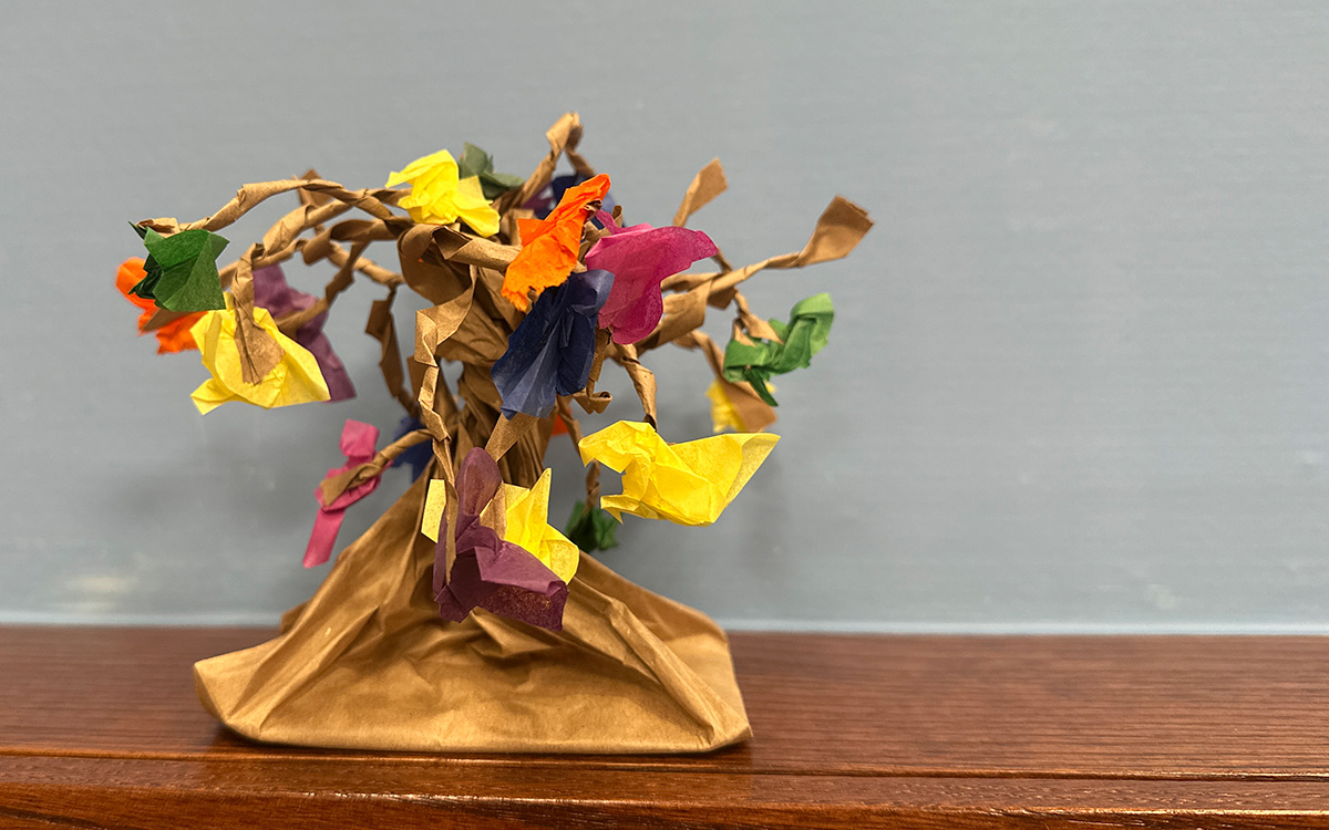 A sculpture -- made of colorful paper and representing a tree-like form -- sits on a wood shelf against a gray background.