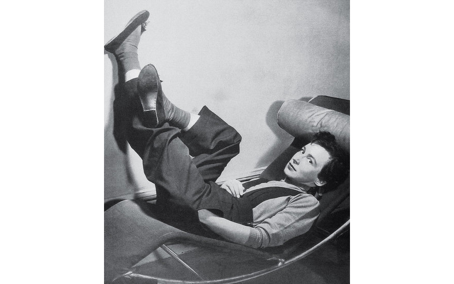Black and white photograph of the woman reclining on a modernist couch with her legs raised up against a wall.