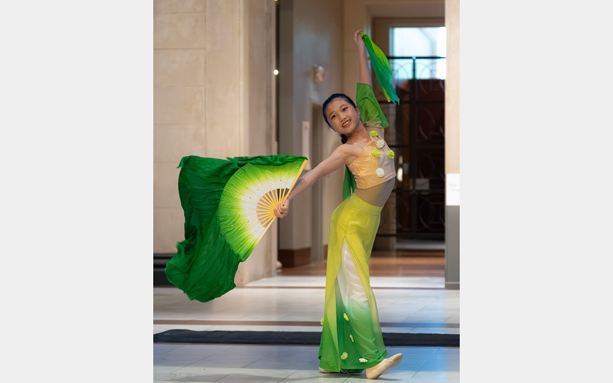 An Asian performer in gorgeous green and yellow attire performs while smiling