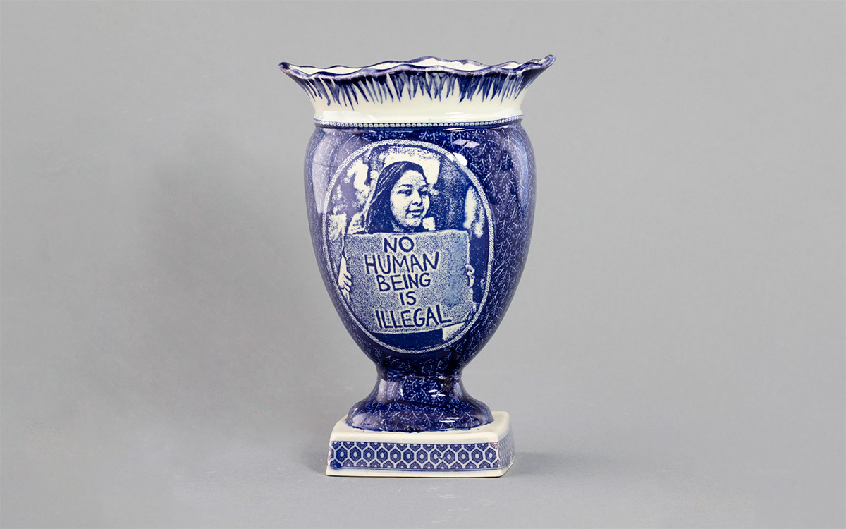 A large vase with a blue transfer of a woman holding a sign that reads "No human being is illegal"