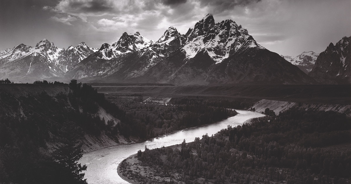 A majestic black and white photo of a jagged mountain range with a curving river in the foreground