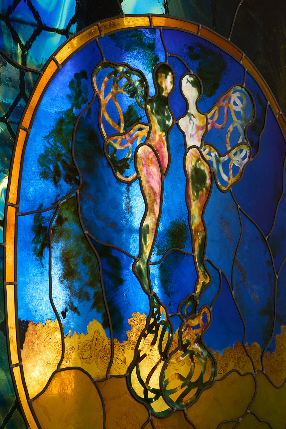 A stained glass window with blue and yellow colors and two feminine figures facing each other above a red flower