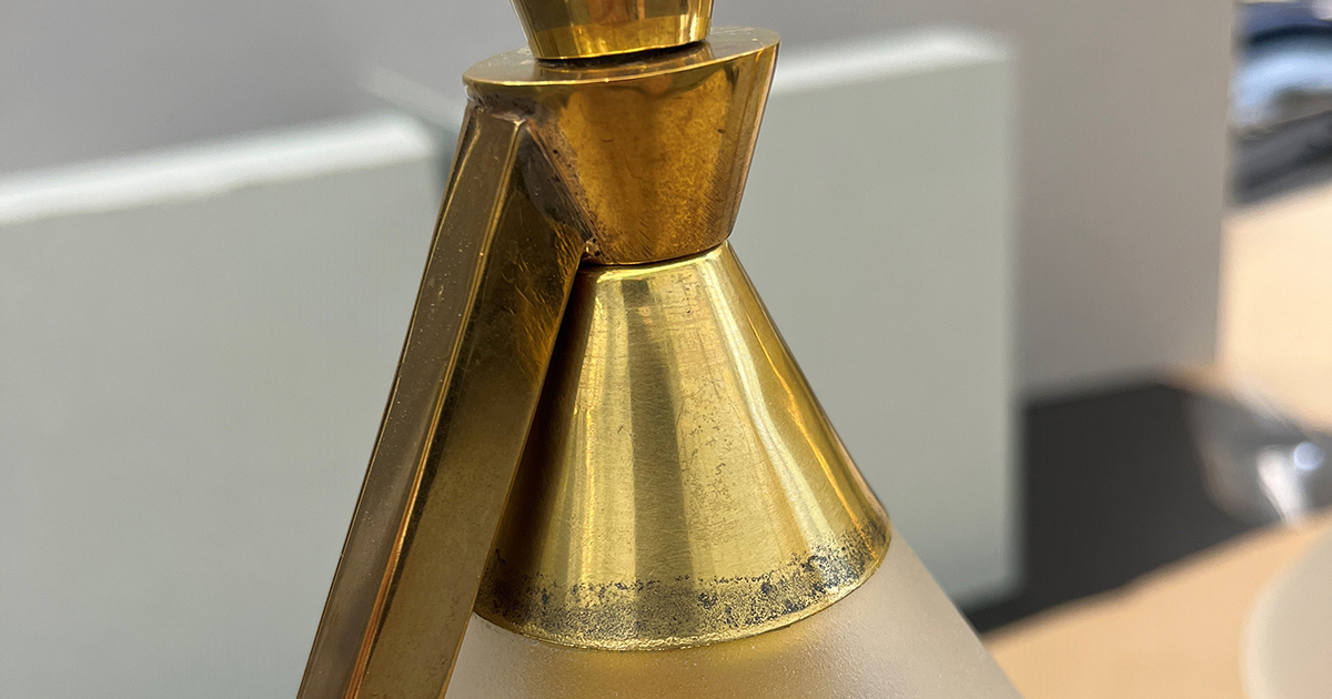 Detail of a gold-colored, angular lamp with dark, speckled tarnish at the bottom of the metal