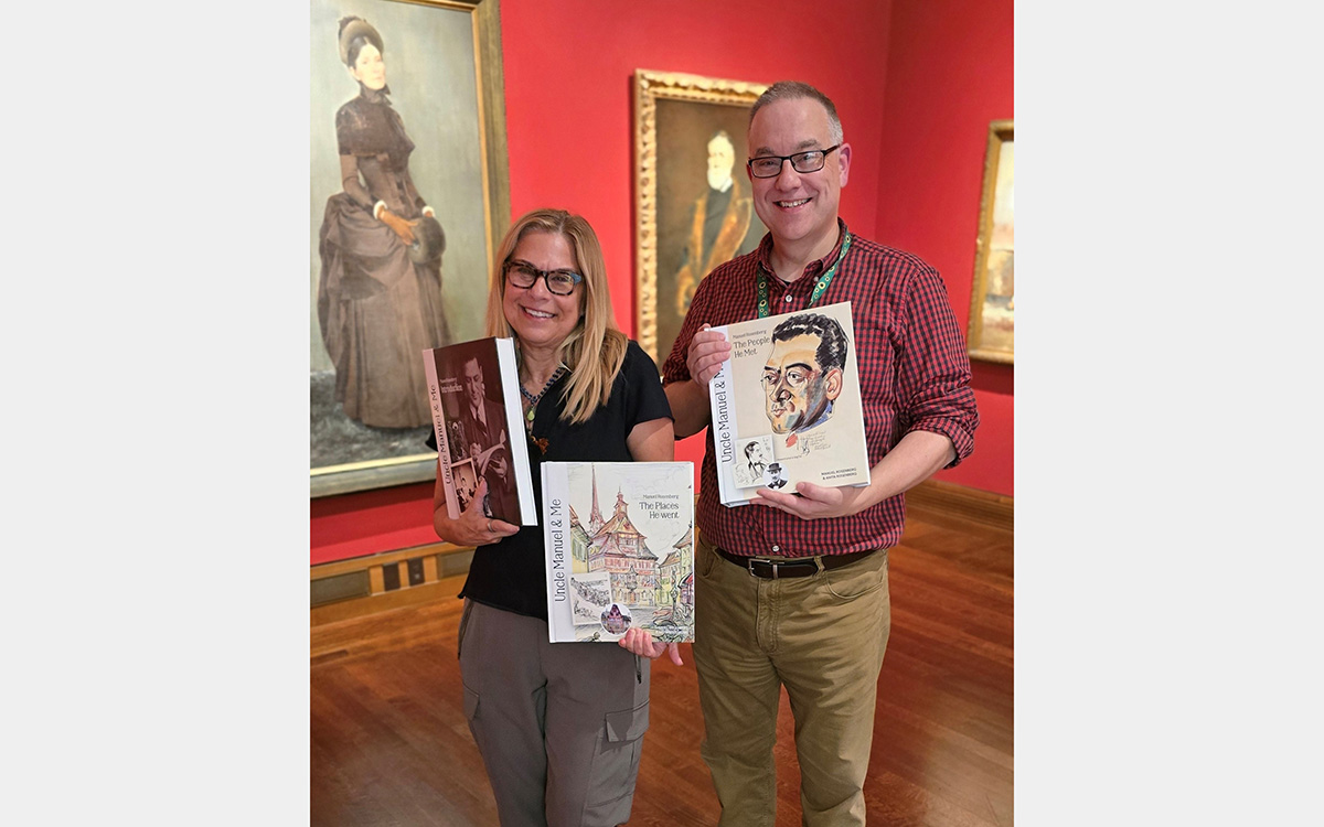A woman and a man hold three books while standing against a red-painted gallery space with large painted portraits in the background.