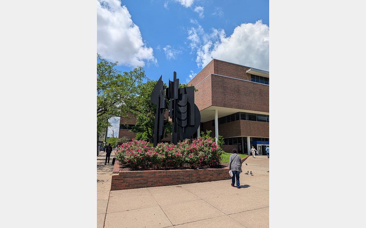 Black metal abstract sculpture standing outdoors surrounded by flowers with a red brick building in the background.