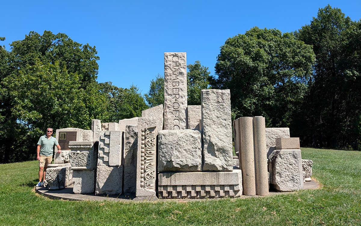Monumental stone blocks in a park-like with green lawn and trees in background. A man stands next to the blocks for scale.