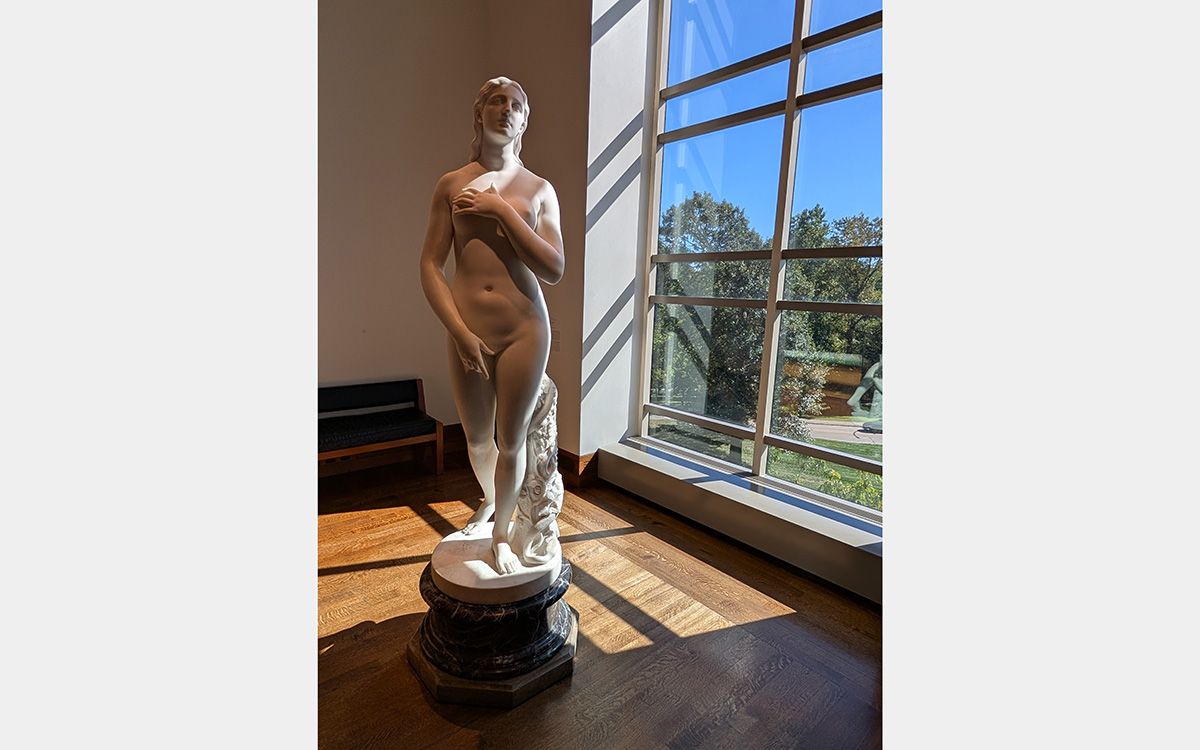 White marble sculpture of a nude woman in front of a floor-length window overlooking greenery and a blue sky.