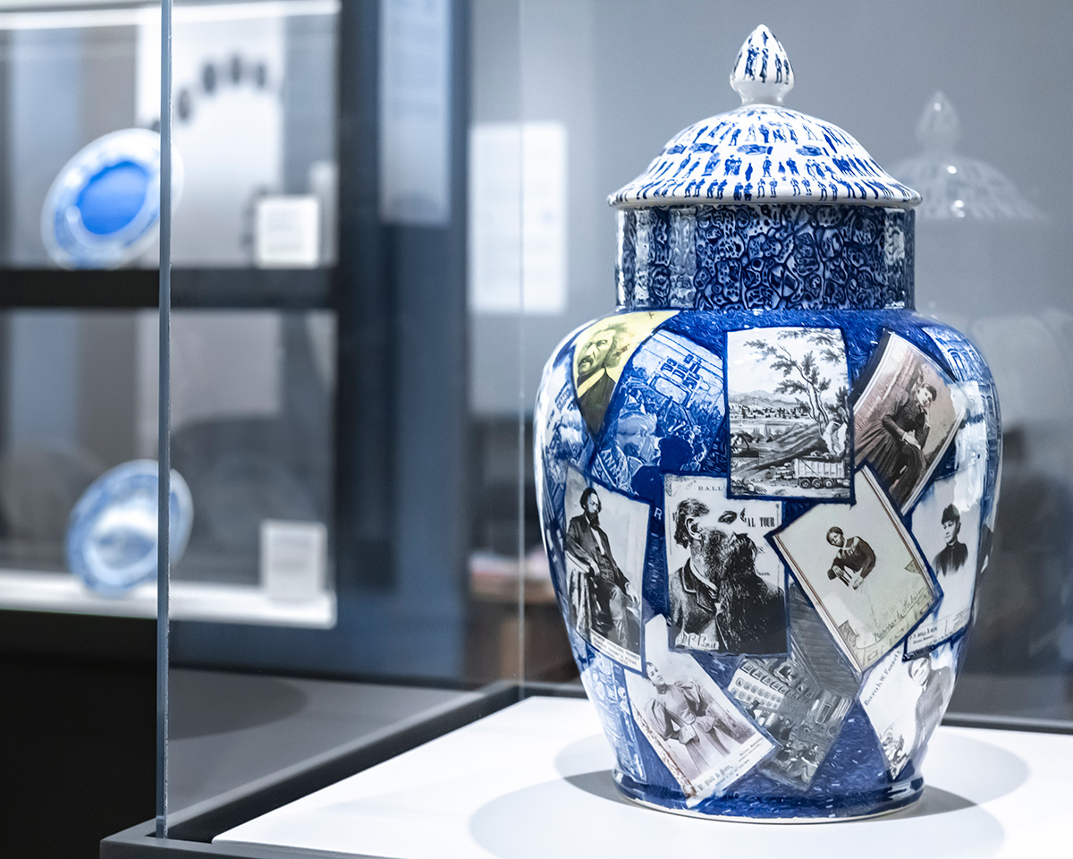 A blue vase decorated with white and black photographic decorations sits within a display case. In the background, two blue and white plates are presented inside another display case.