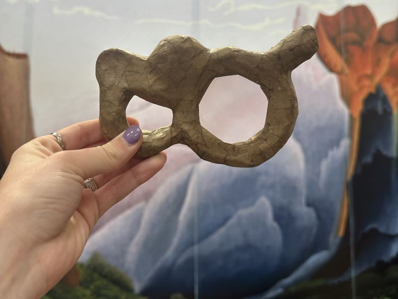 Hand holding magnifying glass framed in paper maché with landscape painting in background