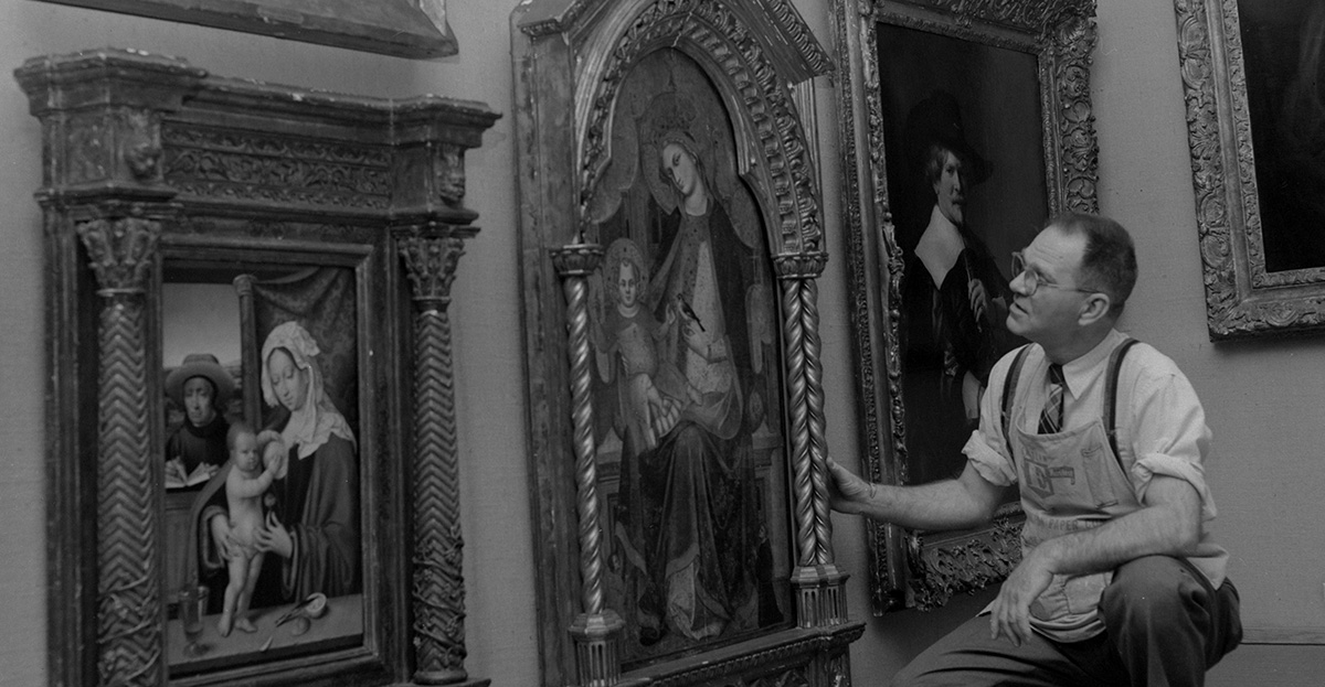 A black and white photo of a white man kneeling and looking up at a painting on a wall