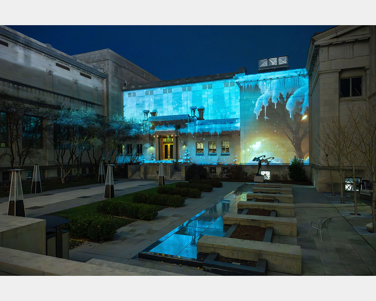 A winter scene in shades of blue is projected onto a wall inside a courtyard of classical architecture built of stone.