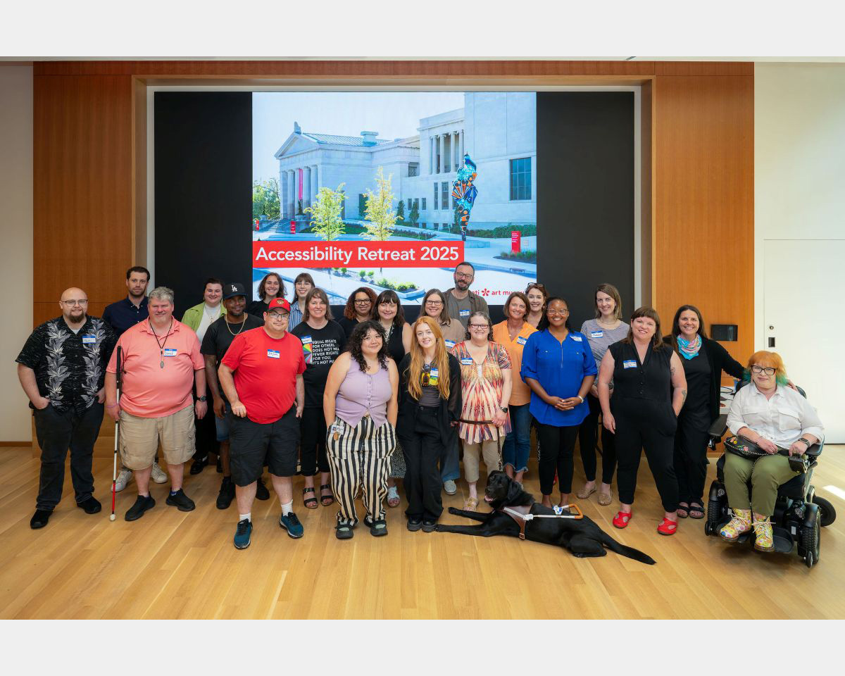 A group of 23 people (one sitting in a wheelchair) and a service dog pose in front of a large project screen stating "Accessibility Retreat 2025"
