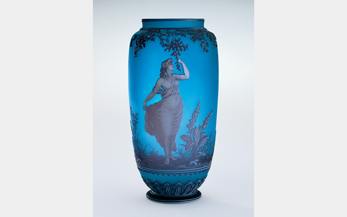 A blue vase with an image of a woman walking through a field (?) while holding a tree branch.