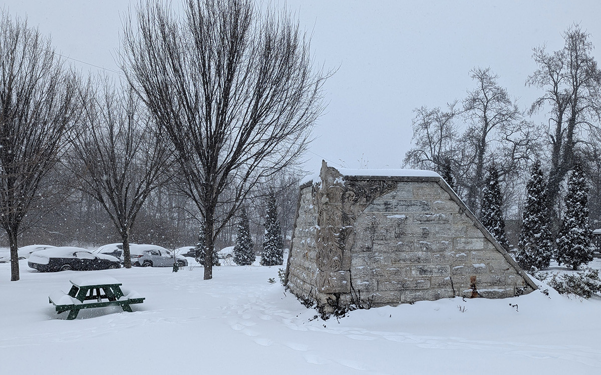 A snow-covered landscape with bare trees, picnic table, and stone structure with an embedded sculptural carving on its corner. Snow carved cars are parked in the distance.
