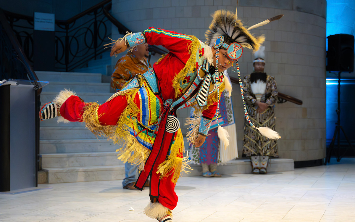 An indigenous dancer in colorful red and yellow attire dances in the Great Hall