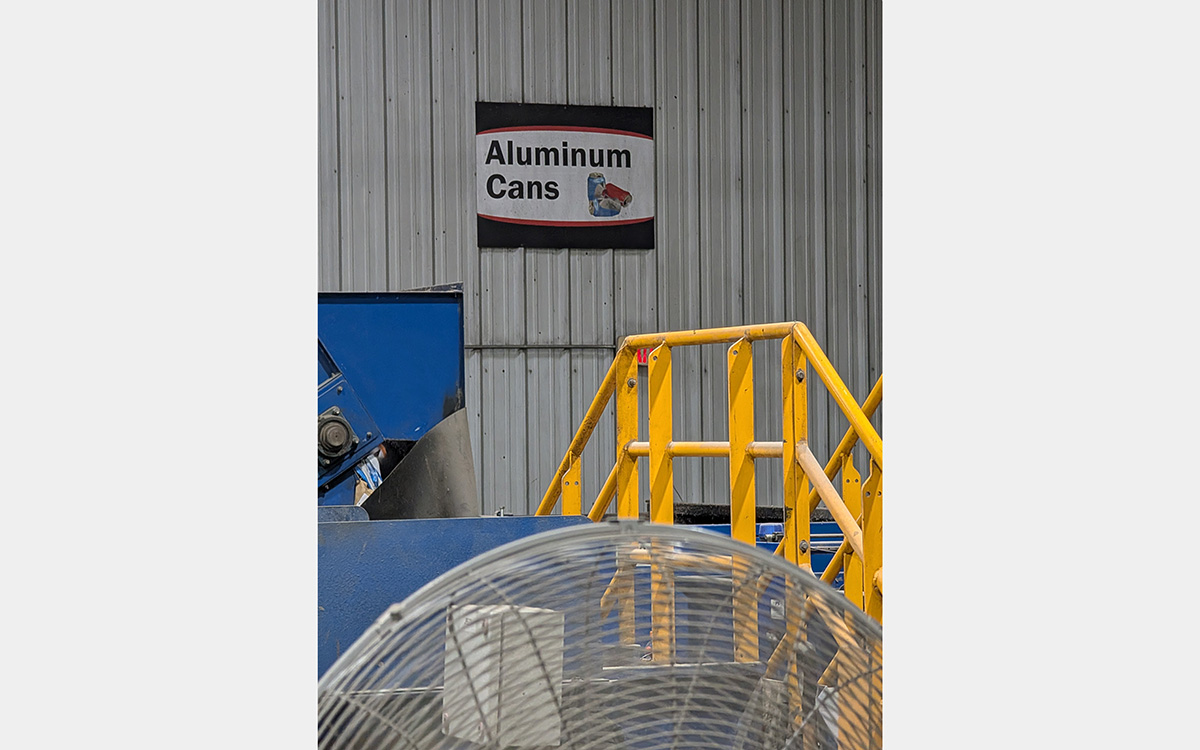 A sign states "Aluminum Cans" inside a warehouse-like space next to a blue steel box and a yellow railing in the foreground