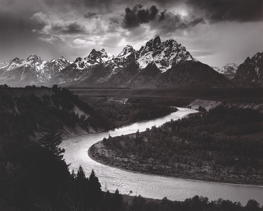 A black and white photograph of a twisting river in front of a jagged mountain range.