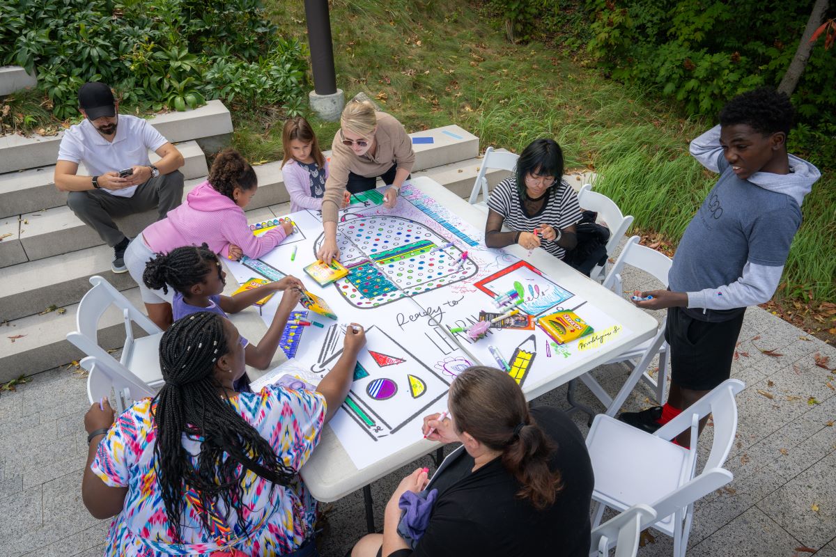 An outdoor view, shown from above, of a square table surrounded by eight people participating in art-making activities.