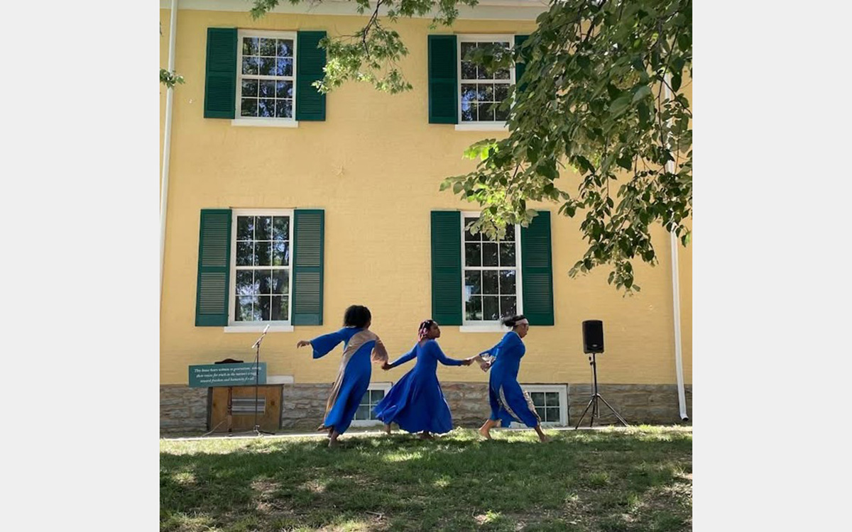 Three girls, dressed in blue, hold hands and dance on a green lawn in front of a yellow house with green shutters.