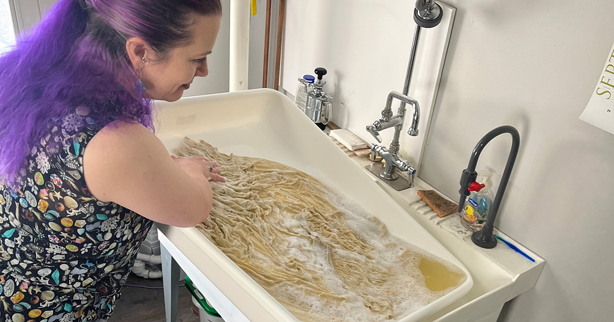 A white woman with purple hair washes the yellowed blouse and skirt in a large utility sink