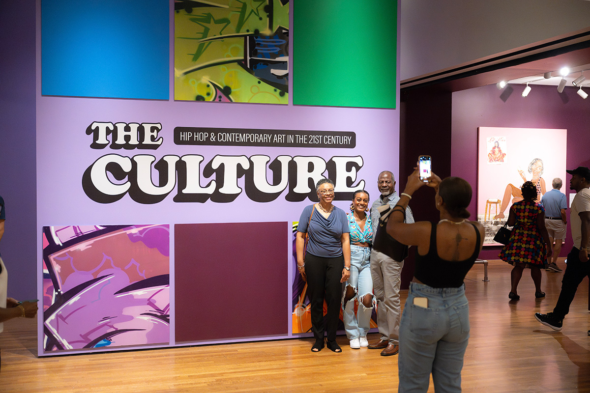 A Black woman takes a picture of her family in front of The Culture title wall